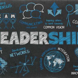 Notes on a blackboard the word "leadership" is highlighted in the center; other terms on the blackboard include: "coaching others", "identifying talent" or "building consensus" some of those terms are held by stick figures or accompanied by symbols like light bulbs or a globe
